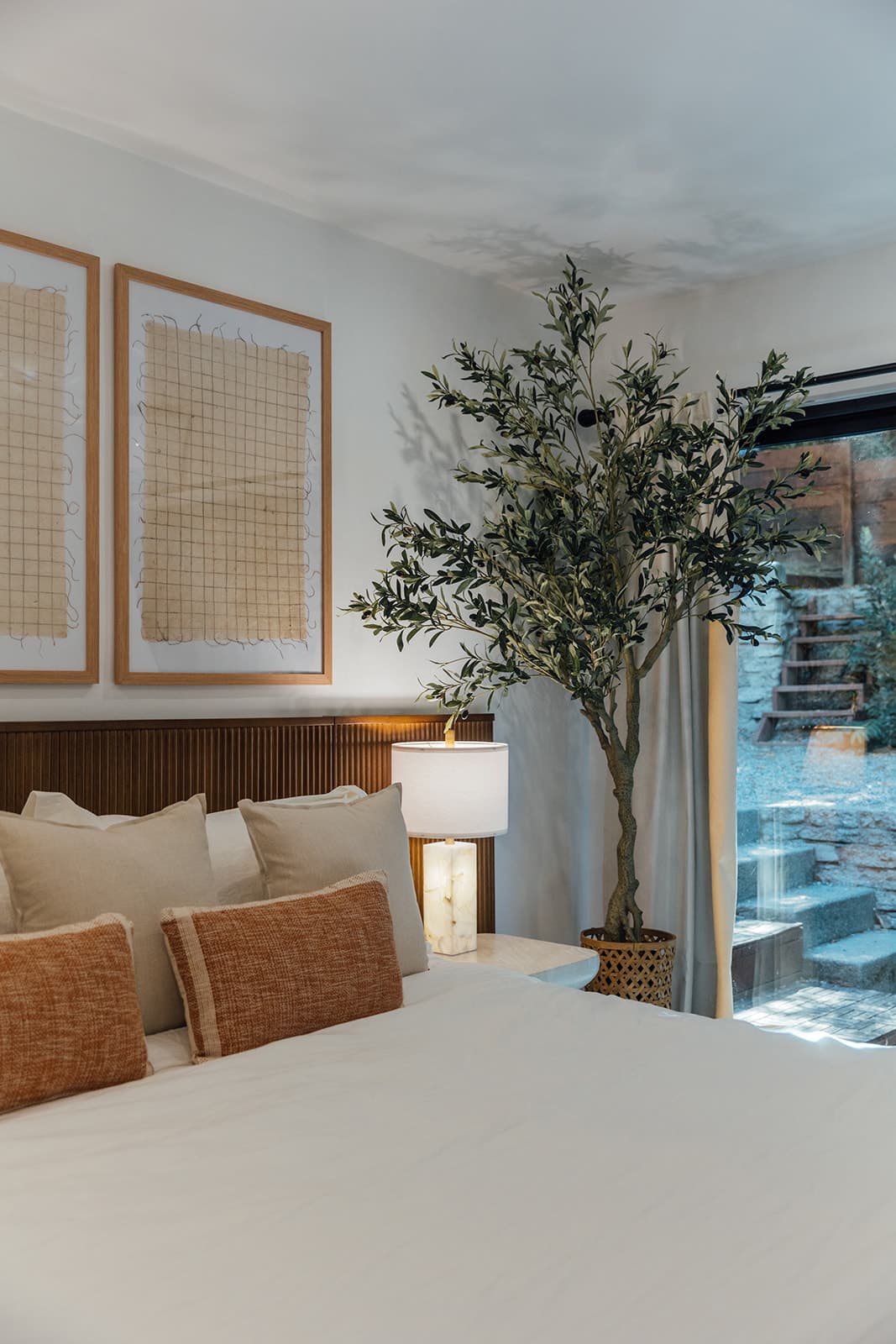 A view across the garden bedroom bed to framed grid textile art, a reeded wood headboard and an olive tree beside glass doors opening to a landscaped stone stairway