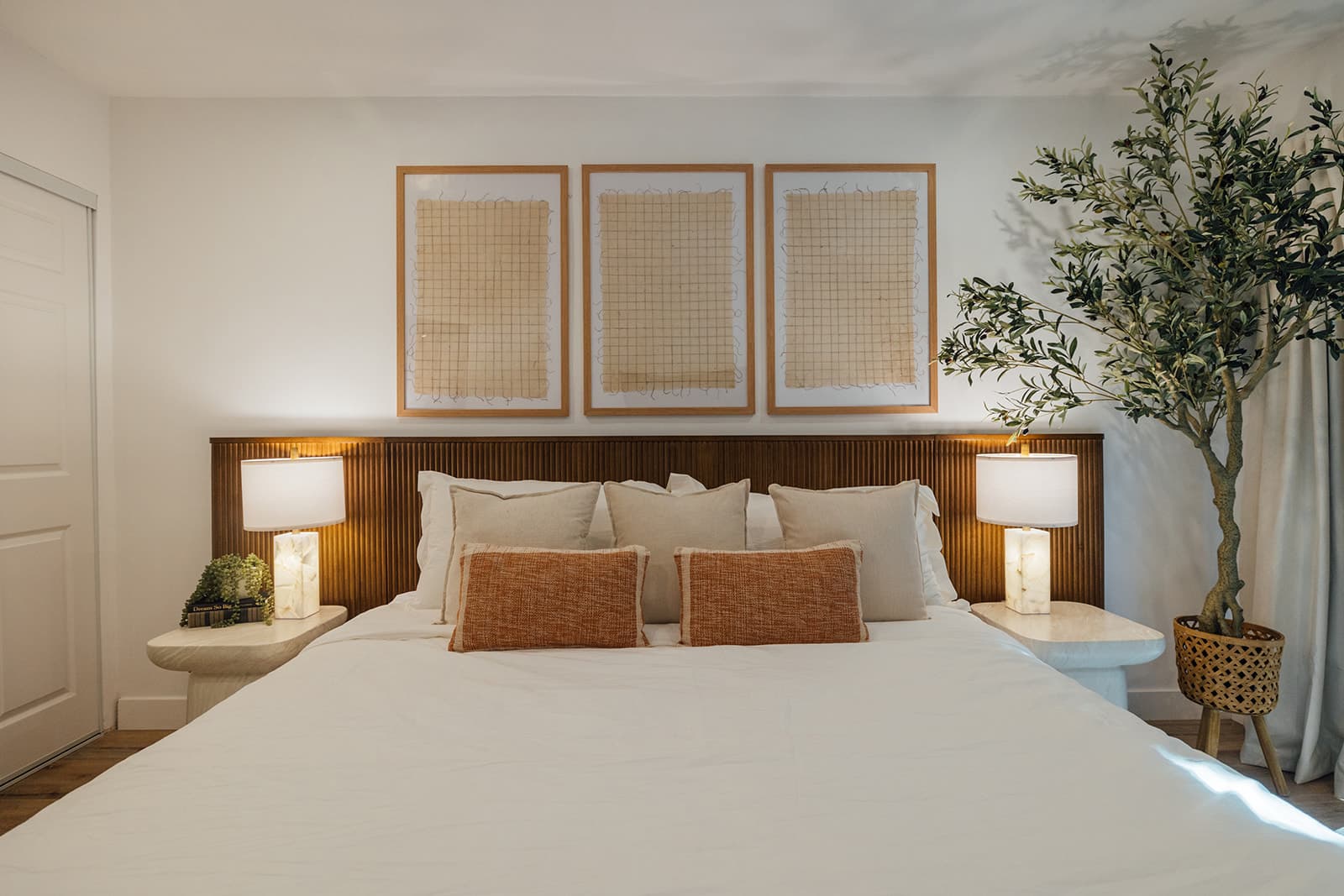 A king bed with a fluted wood headboard, alabaster lamps on curved nightstands, three framed grid prints and an olive tree at the right