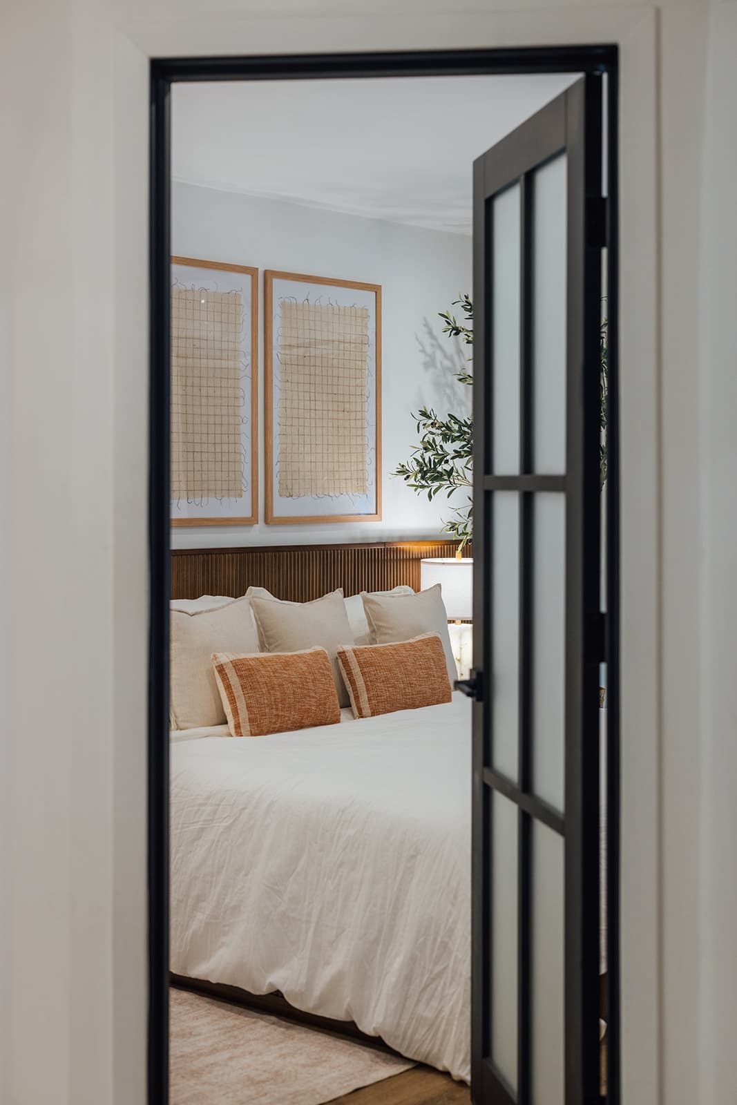 A king guest bedroom seen through its glass door, dressed with rust lumbar pillows