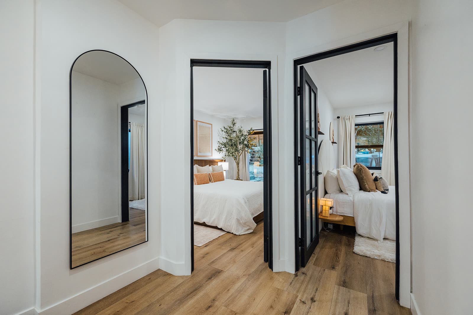 A hallway with an arched full-length mirror opening onto two guest bedrooms — the garden bedroom with its olive tree at left, the cozy king at right