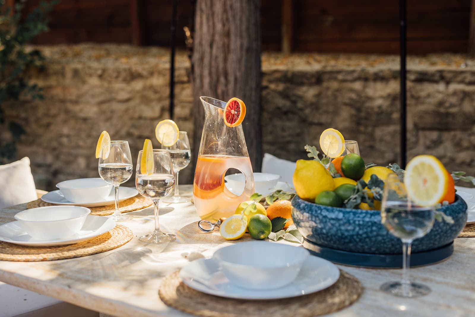 An outdoor travertine table set with white bowls, lemon-garnished wine glasses, a pitcher of blood-orange drink and a bowl of citrus against a stone wall