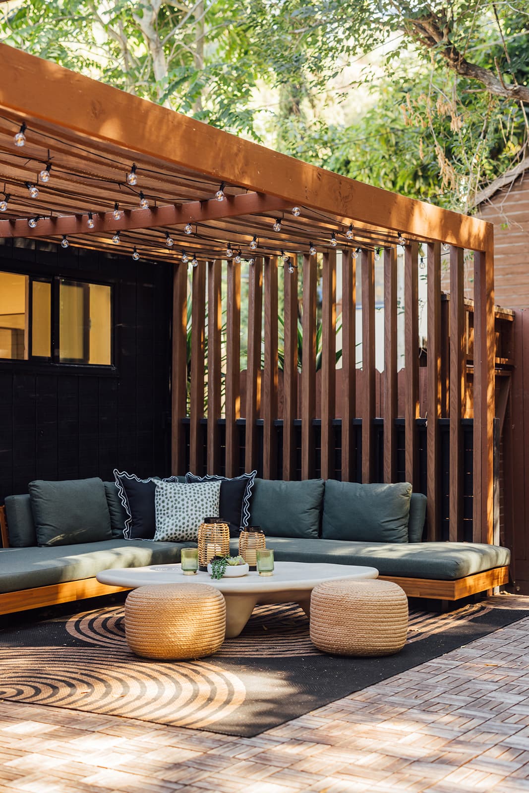 A green-cushioned corner sectional under a wood-slat pergola strung with cafe lights, two woven poufs and a round coffee table on a patterned rug over herringbone tiles