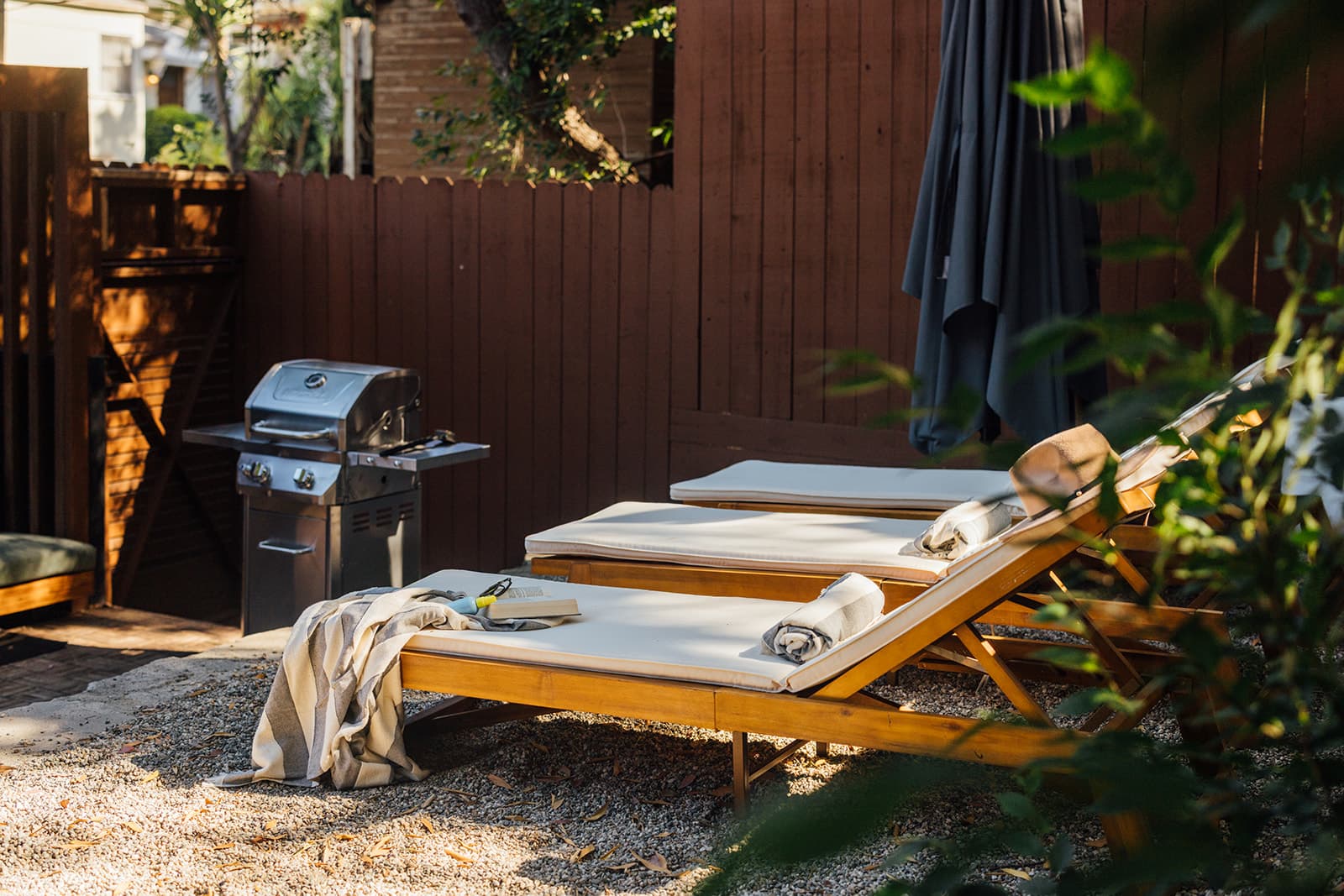 Two wooden chaise loungers with cream cushions and rolled towels on gravel beside a stainless steel gas grill against a dark wood fence