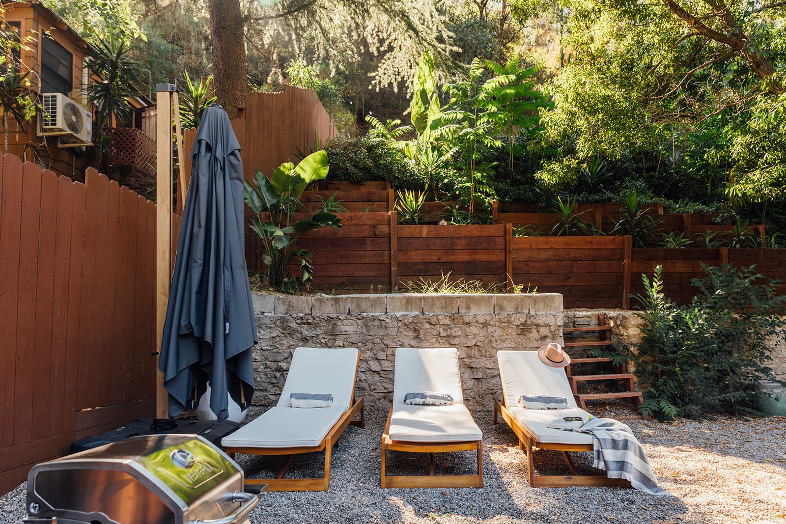 Three wood-framed sun loungers with cream cushions along a stone retaining wall backed by tiered planters and greenery, a closed umbrella and covered grill to the left