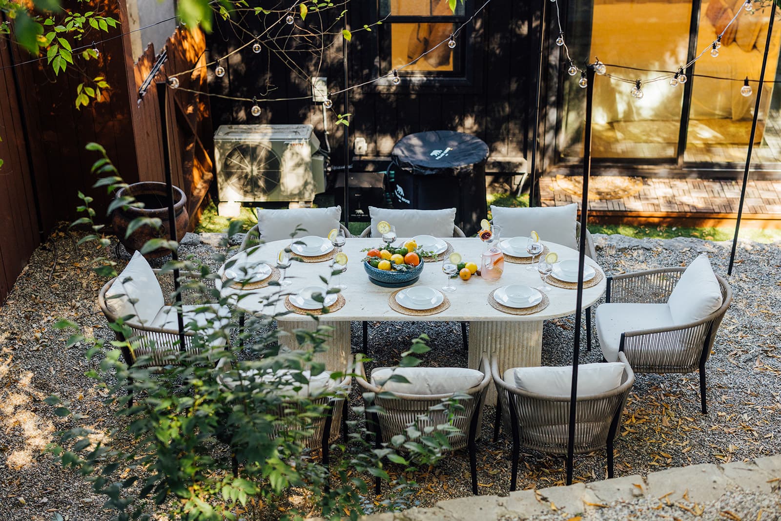 An oval travertine table set for six with white plates, woven placemats and a bowl of citrus, cushioned rope chairs on gravel, string lights and the board-and-batten wall behind
