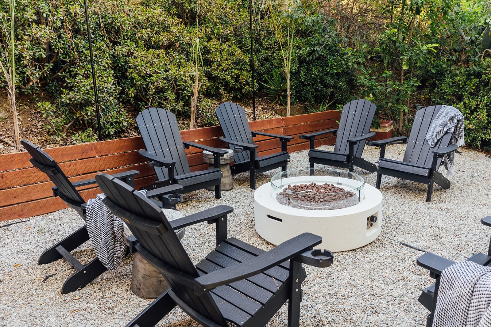 Six black Adirondack chairs with woven blankets encircling a round white gas fire pit filled with lava rock behind a glass wind guard, on a gravel patio backed by greenery