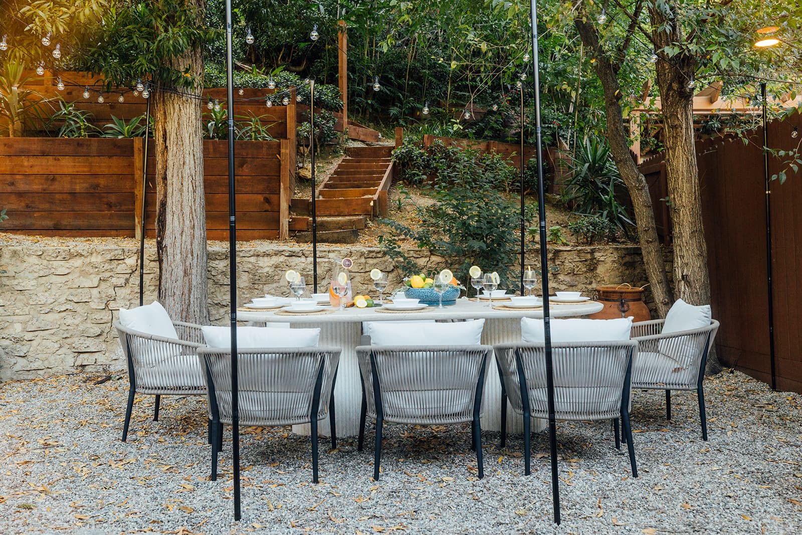 An oval stone dining table set with citrus-garnished glasses and a fruit bowl, surrounded by grey rope armchairs on gravel beneath trees and a tiered wood retaining wall with steps