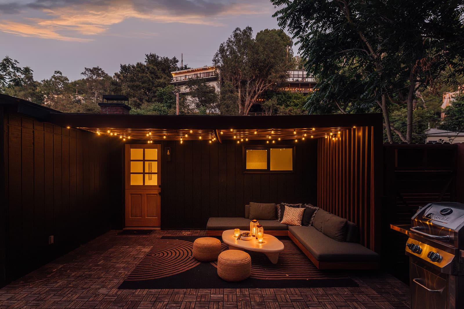 The black board-and-batten house at dusk, string lights over the terrace lounge and a warm-lit Dutch door