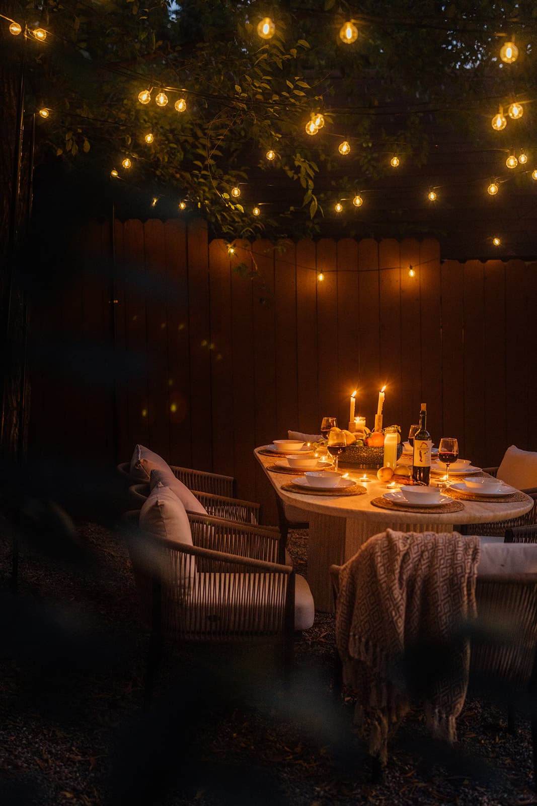 A candlelit dining table set with white bowls and taper candles against a wood fence, cushioned rope chairs and a fringed throw, under strands of string lights