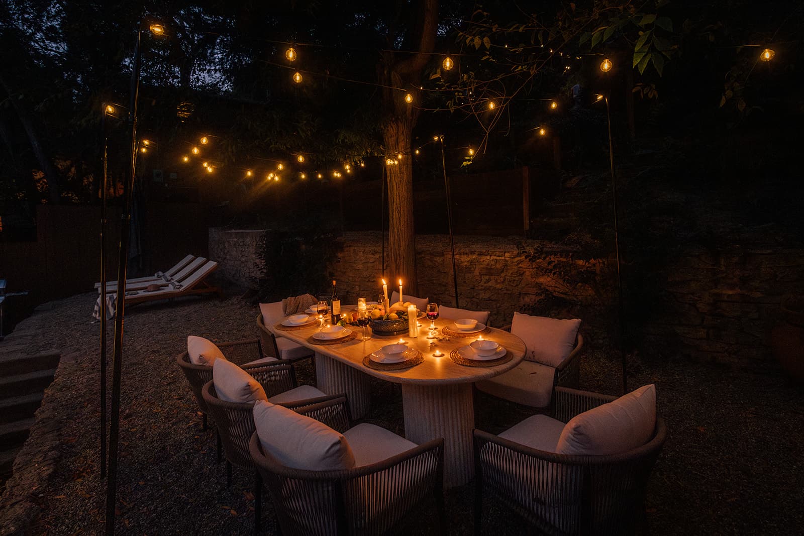 An oval table lit by candles and set for six with rope chairs on a gravel patio beside a stone wall, two wooden loungers to the left, string lights strung from a central tree