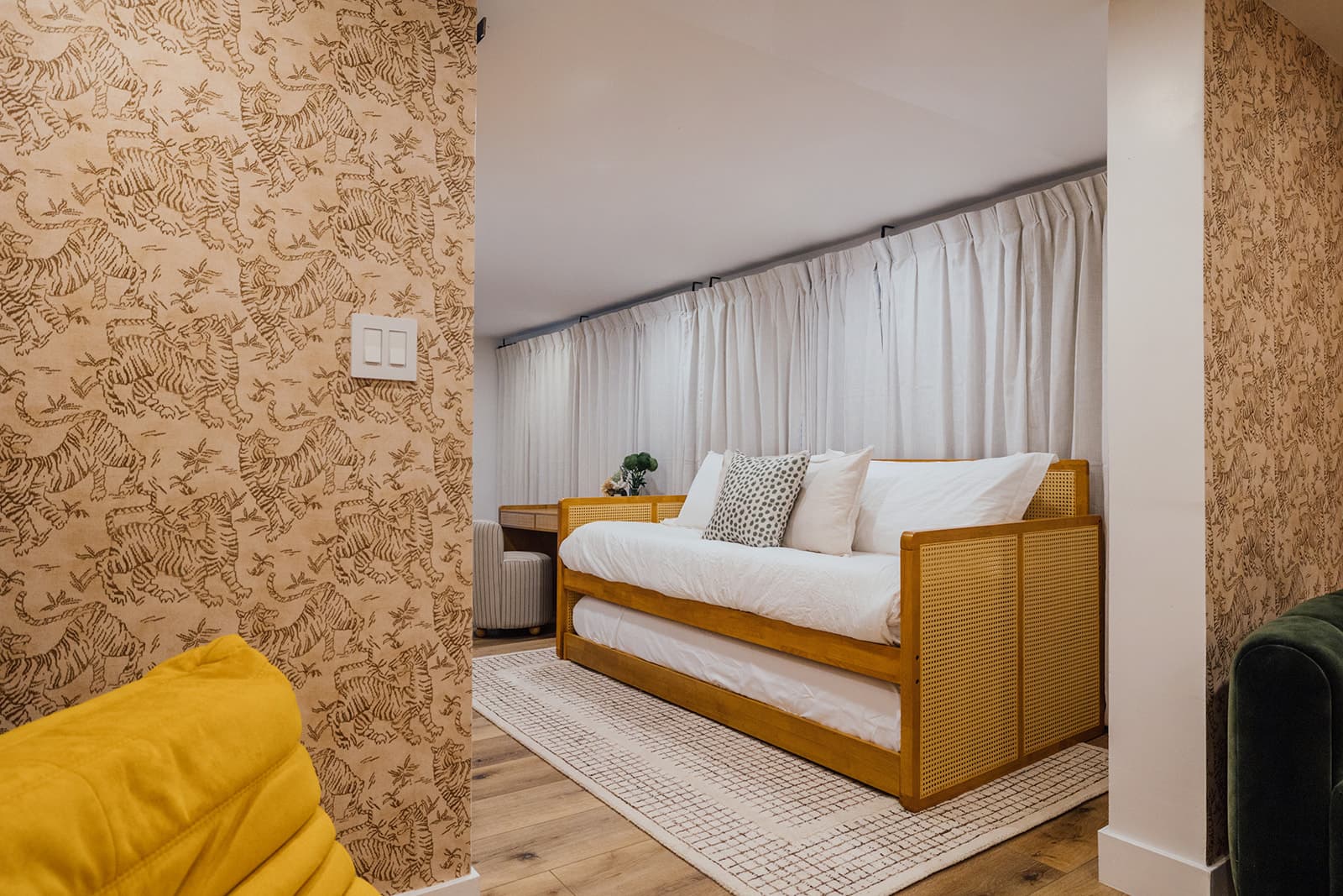 A wood-framed cane daybed with a pull-out trundle made up in white bedding on a woven rug, framed by tiger-print walls and the edge of a yellow Togo chair