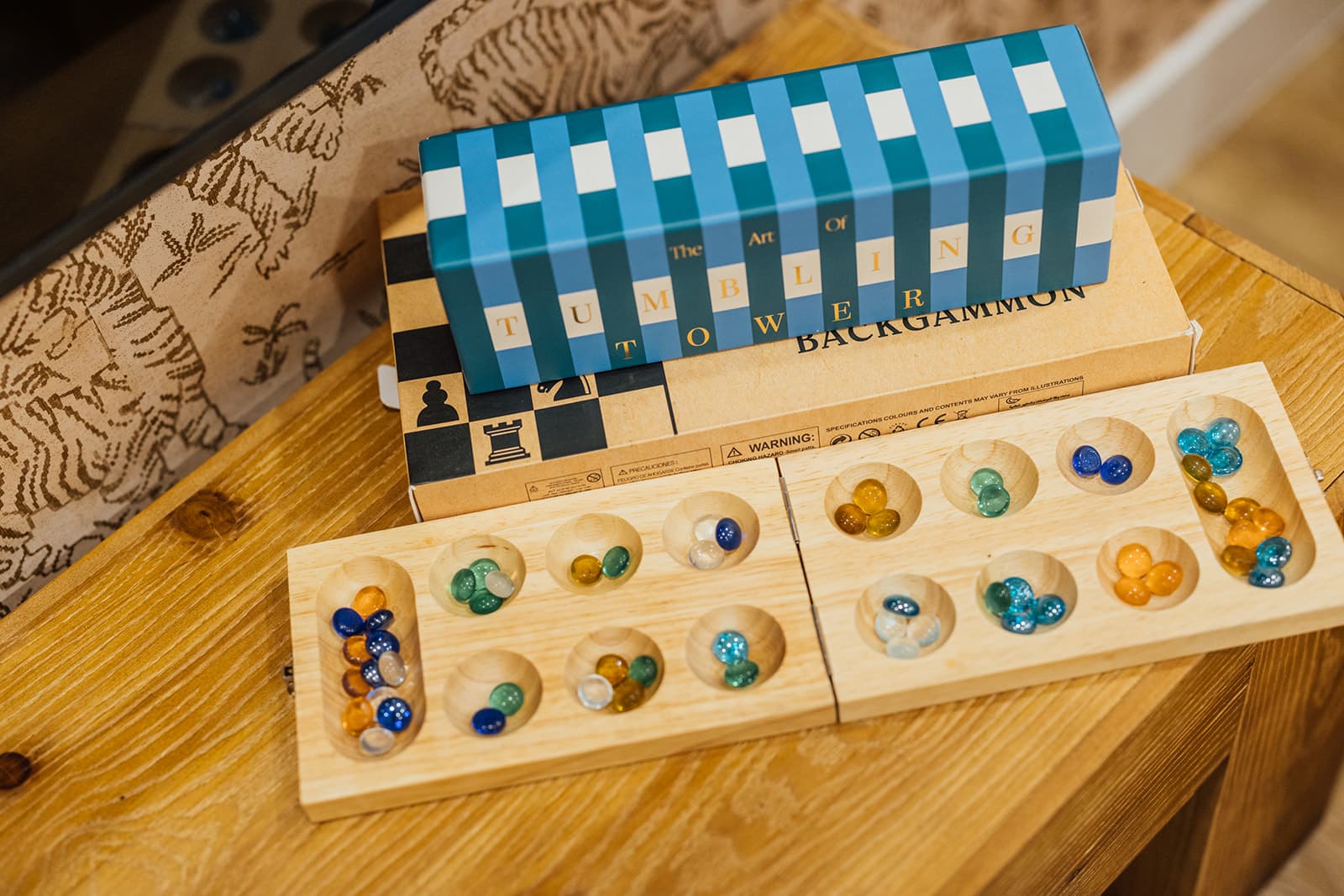 A wooden mancala board mid-game with colored glass stones, topped by a striped tumbling-tower box and a backgammon set, on a light-wood table