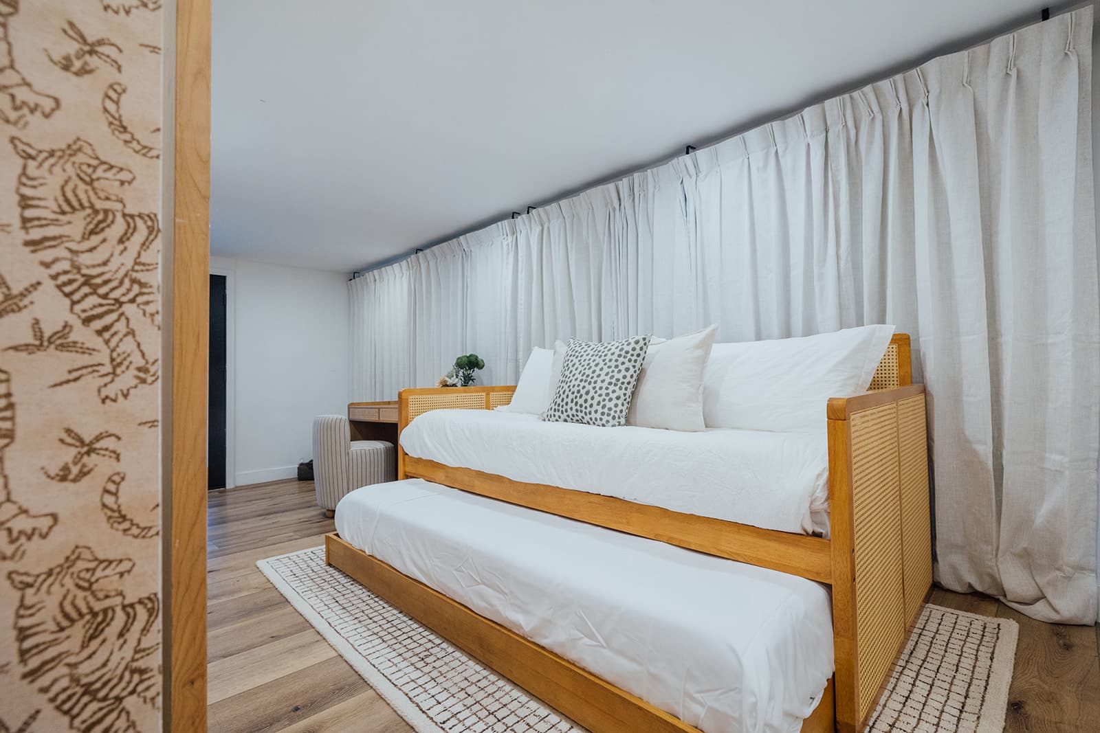 A cane daybed made up with white bedding and a pull-out trundle beneath, against floor-length linen curtains and tiger-print wallpaper