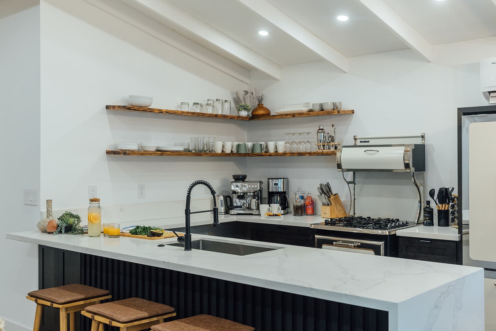 A marble peninsula and range with burnt-wood floating shelves, a black gooseneck faucet and coffee appliances against a bright white wall