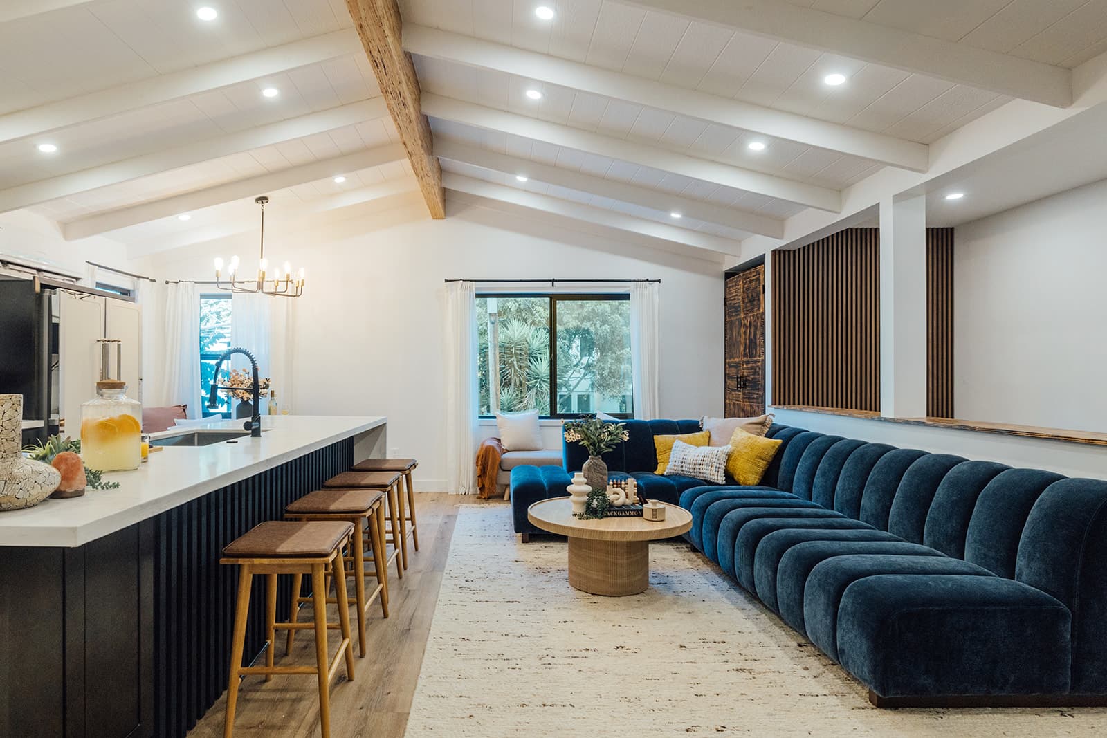 The great room under vaulted, whitewashed beams: the marble island with wooden bar stools and the long blue velvet sectional