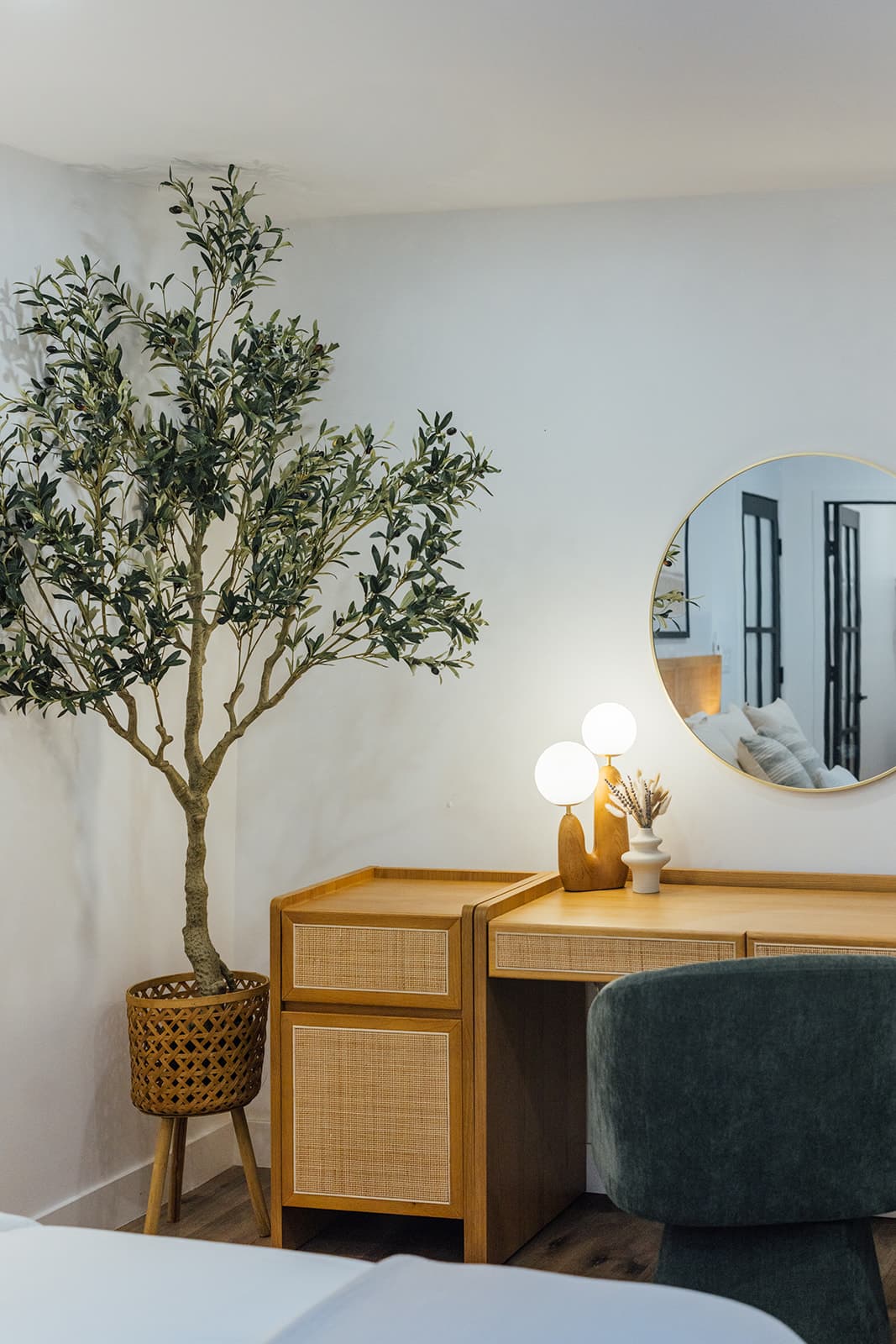 A wood-and-cane vanity with a green velvet stool, twin globe lamp and round gilt mirror beside a potted olive tree