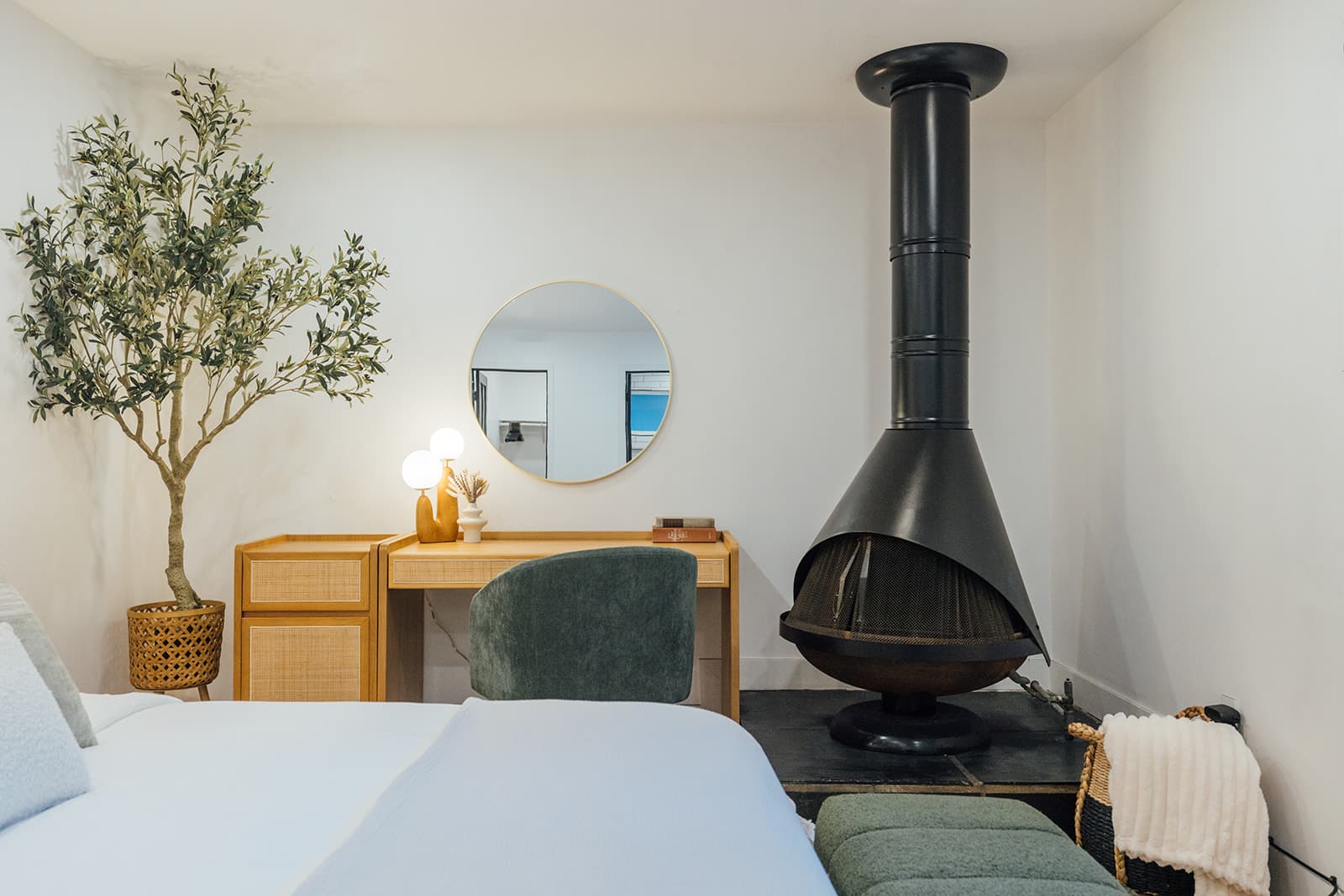 A black cone-shaped freestanding fireplace with a tall flue in the corner beside the cane vanity, round mirror and olive tree, the bed in the foreground